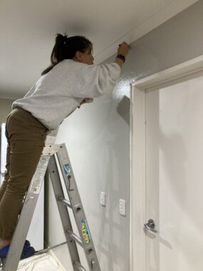 A middle-aged woman stands on a ladder and uses a small brush to paint the upper edge of a wall, where it meets the crown moulding. The ceiling and a door to her right are white, while she's painting the wall a light gray.