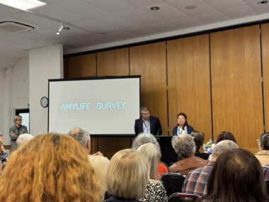 Two people, a woman and a man, stand behind a podium at the front of a room. There's a large screen to their right that reads "Amylife survey." The photo is taken from the audience, several rows back.