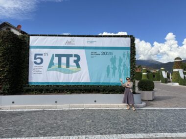 A woman stands in front of a teal blue and white billboard that says 5th ATTR 2025 (there are other words, but they are too small to see). Behind the sign is a picturesque setting of the Italian countryside. 
