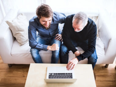 Two men on a couch look at a laptop on top a coffee table.